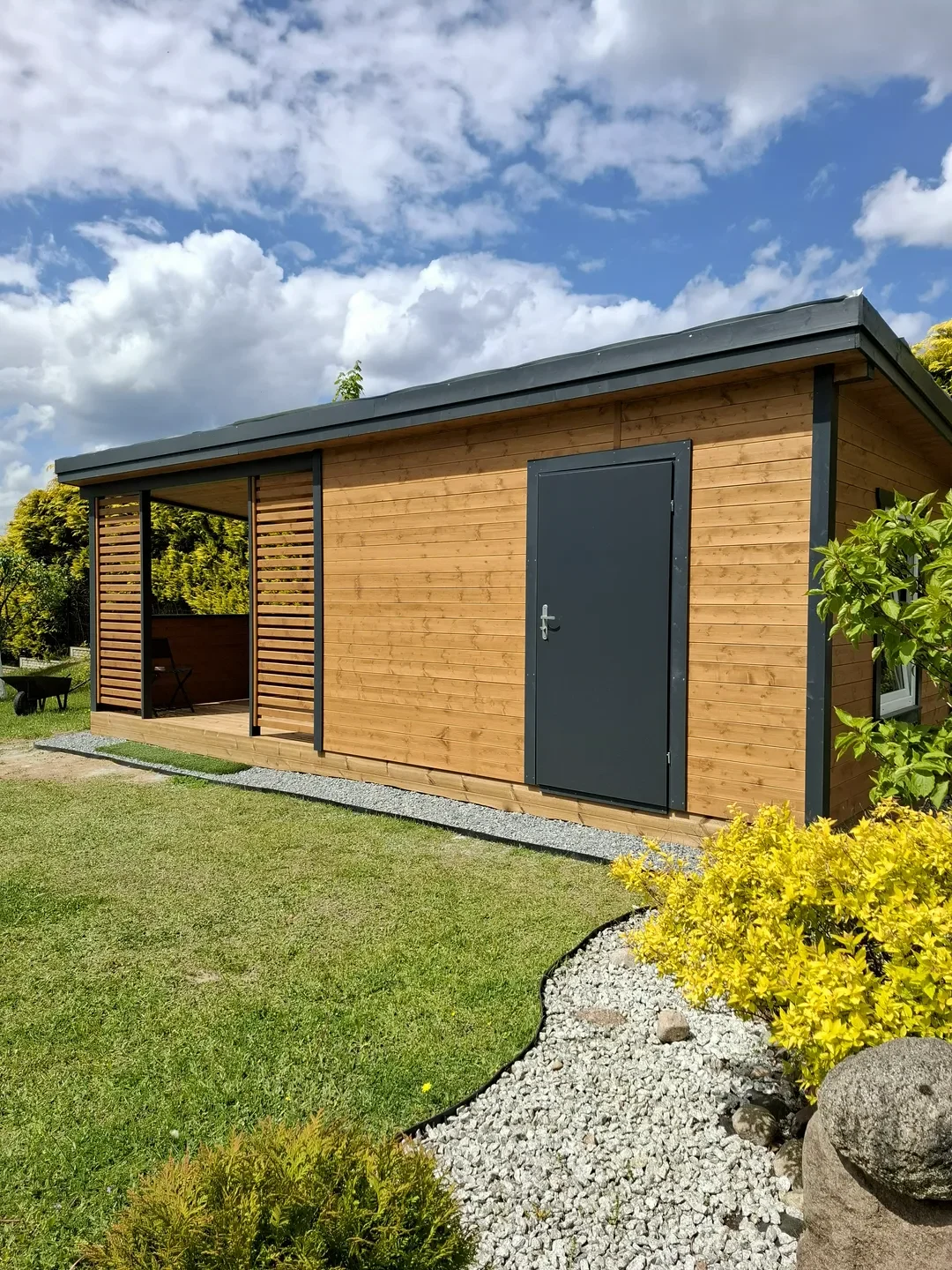 Drewniany domek ogrodowy z tarasem, metalowymi drzwiami i ozdobnym żwirem na trawniku