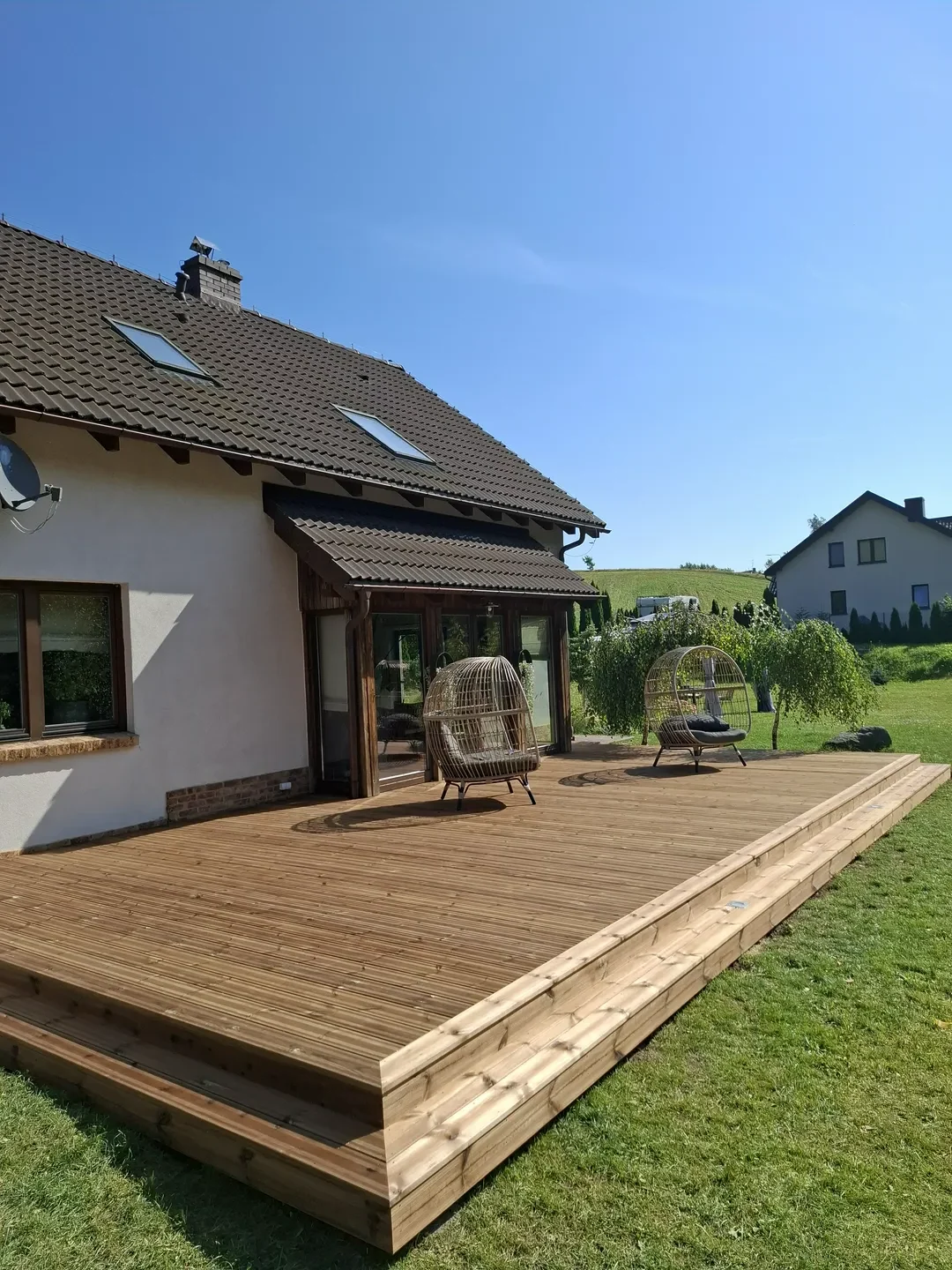 Taras drewniany przy domu z dwoma wiszącymi fotelami i widokiem na zielony ogród