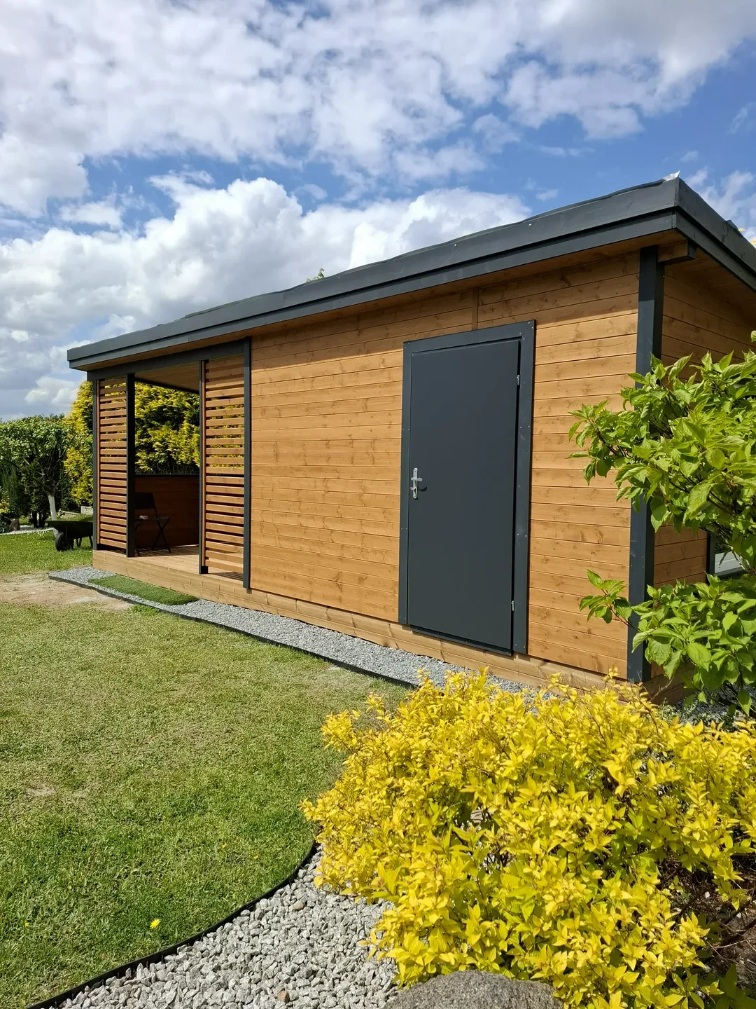 Domki narzędziowe drewniane z szarą metalową furtką i ozdobnymi żółtymi krzewami przy trawniku