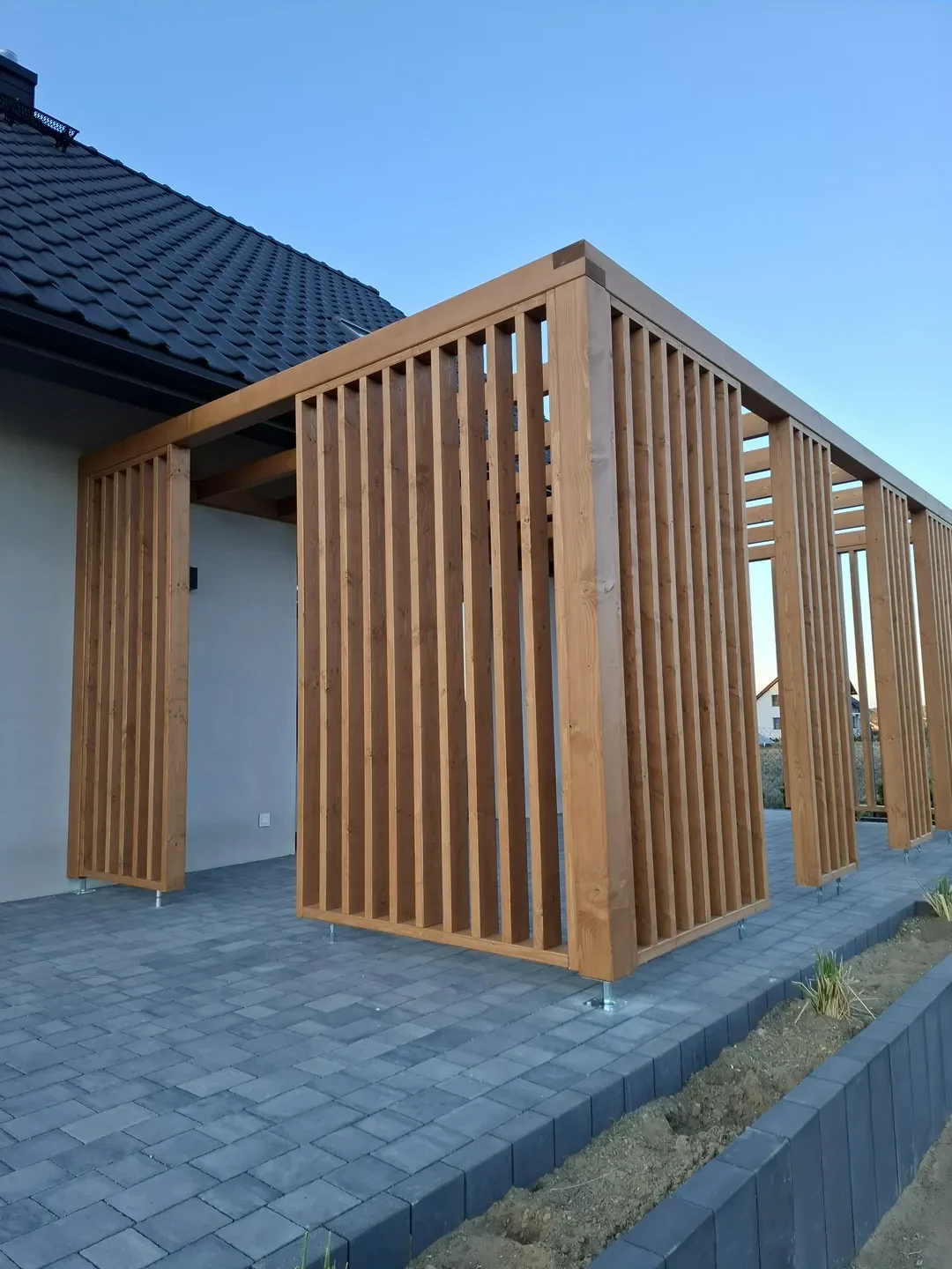 Pergole drewniane z pionowymi lamelami tworzące nowoczesne zadaszenie tarasu przy budynku mieszkalnym