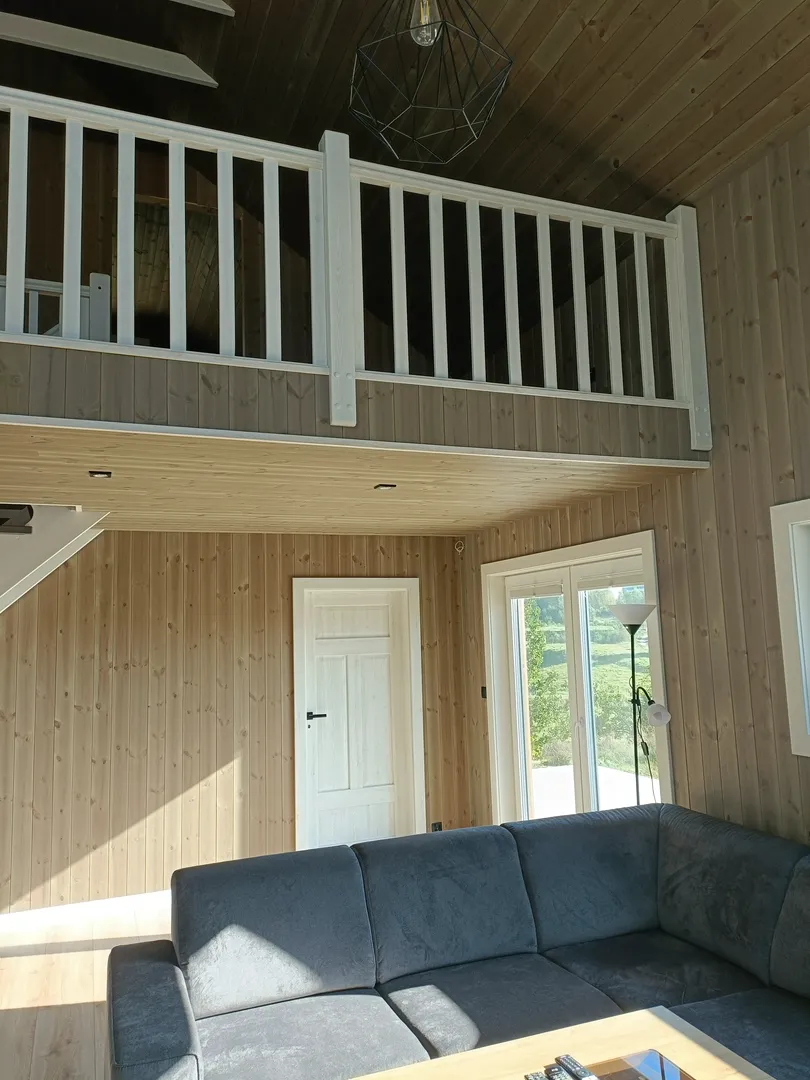 Wykończenia wnętrz drewnianymi panelami: salon z antresolą, białymi drzwiami i szarą sofą