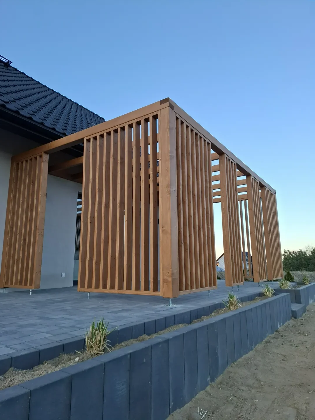 Pergole ogrodowe lekka drewniana konstrukcja z pionowymi listwami osłonowymi przy tarasie z kostki