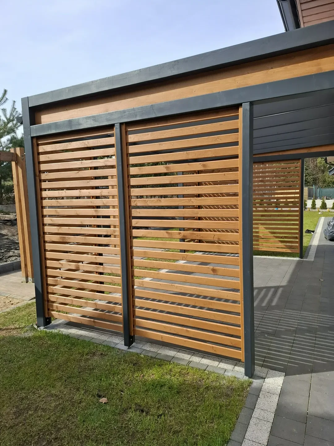 Drewniana pergola ogrodowa z poziomymi deskami i stalową ramą na tle trawnika i kostki brukowej