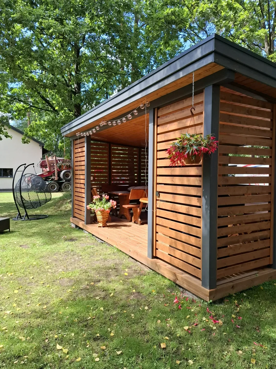 Drewniana altana ogrodowa z tarasem, wiszącą donicą i dekoracyjnymi lampkami