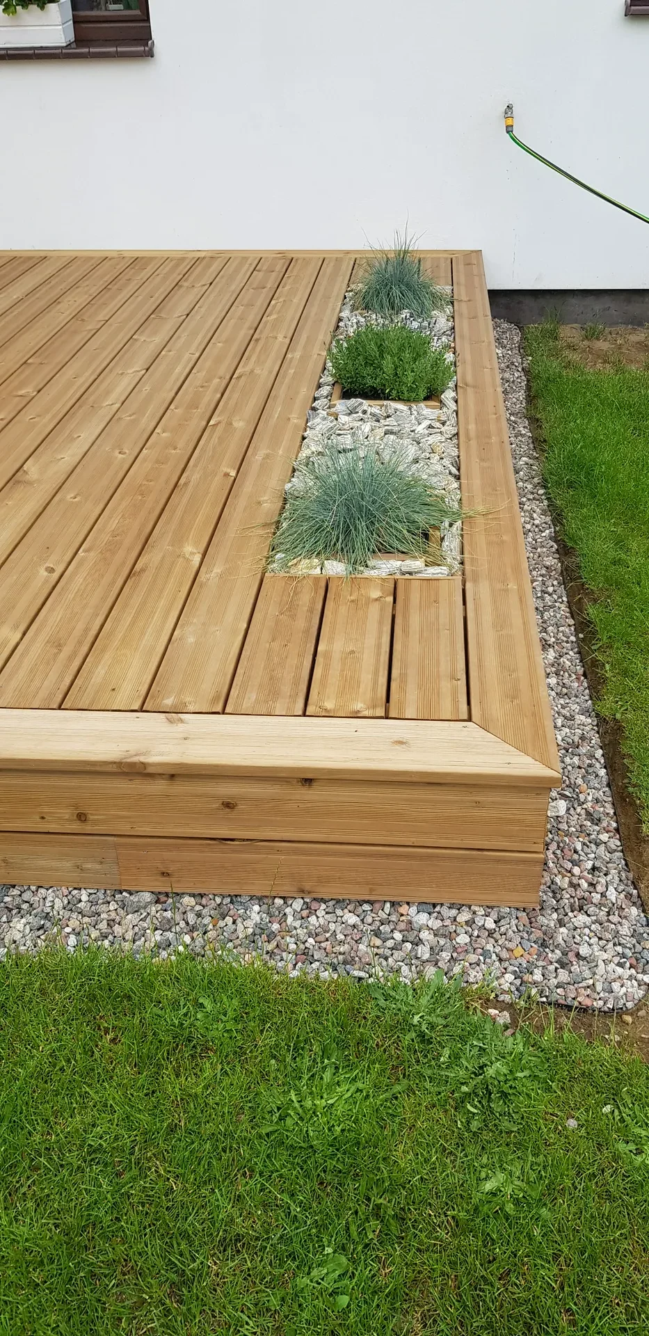 Taras z drewnianych desek z wbudowaną donicą z roślinami i obsypką z kamieni przy trawniku