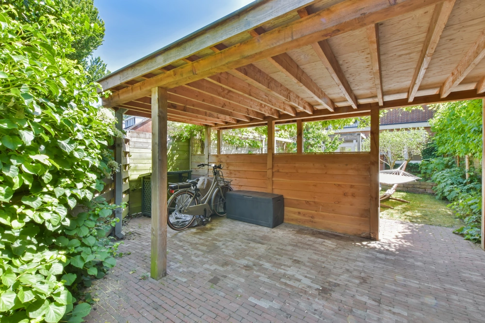 Drewniana wiata garażowa na utwardzonym podjeździe z rowerem i skrzynią, w tle ogród i hamak